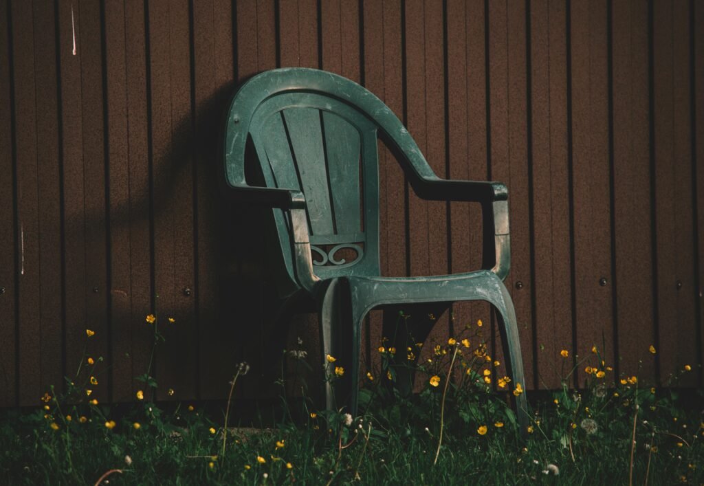 An outdoor plastic chair placed against a wooden wall surrounded by wildflowers in low light.